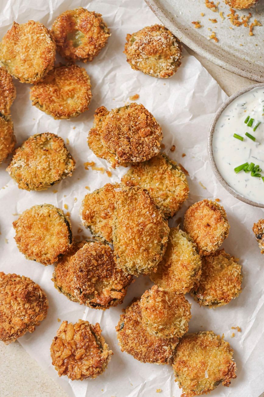 Golden breaded pickle chips on parchment with a bowl of ranch.