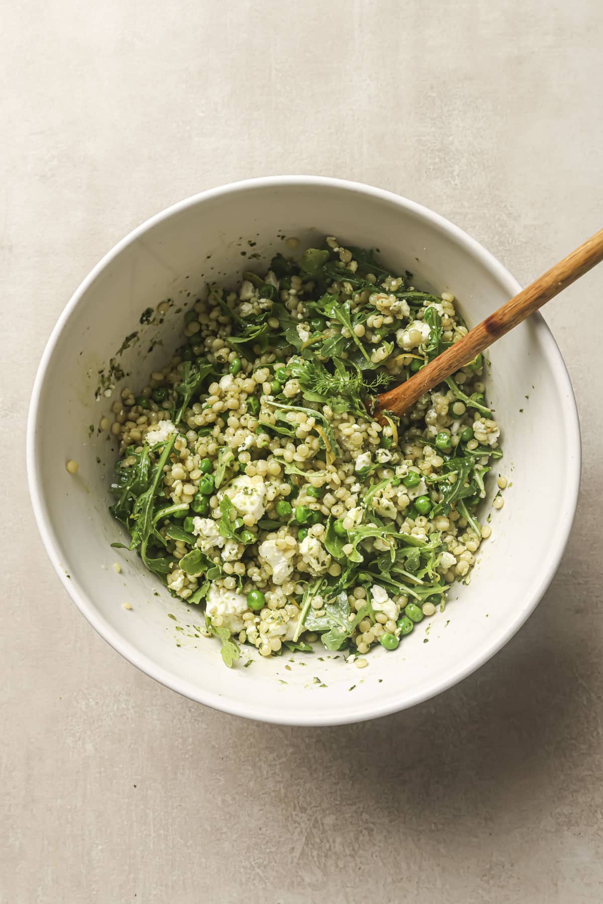 Stirring couscous with arugula, feta, lemon juice and pesto in white bowl.