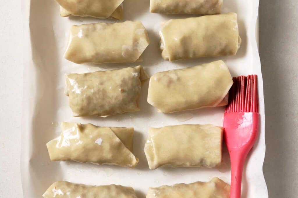 Wrapped cheeseburger egg rolls on a piece of parchment with a silicon brush.