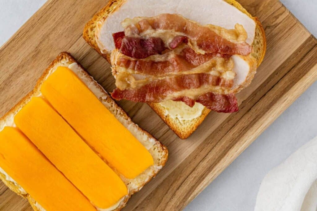 Adding bacon, turkey, and cheddar to two slices of bread.