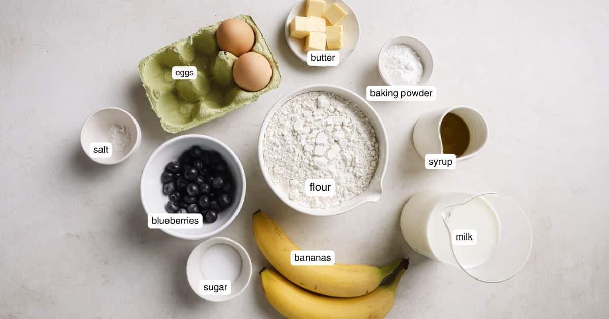 Two bananas near bowls of blueberries, flour, milk and two eggs.