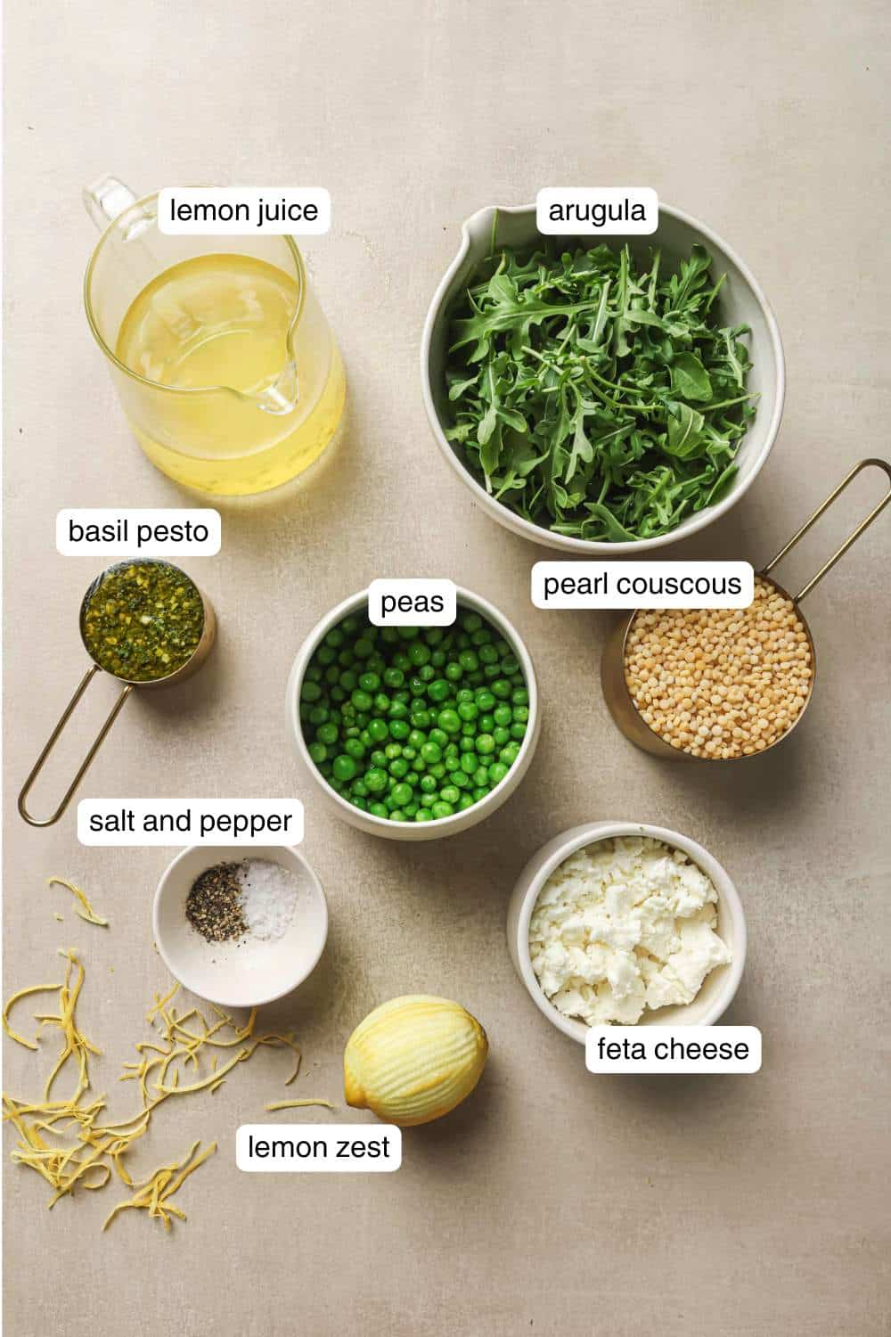 Bowls with arugula, uncooked couscous, feta cheese and peas near a zested lemon.