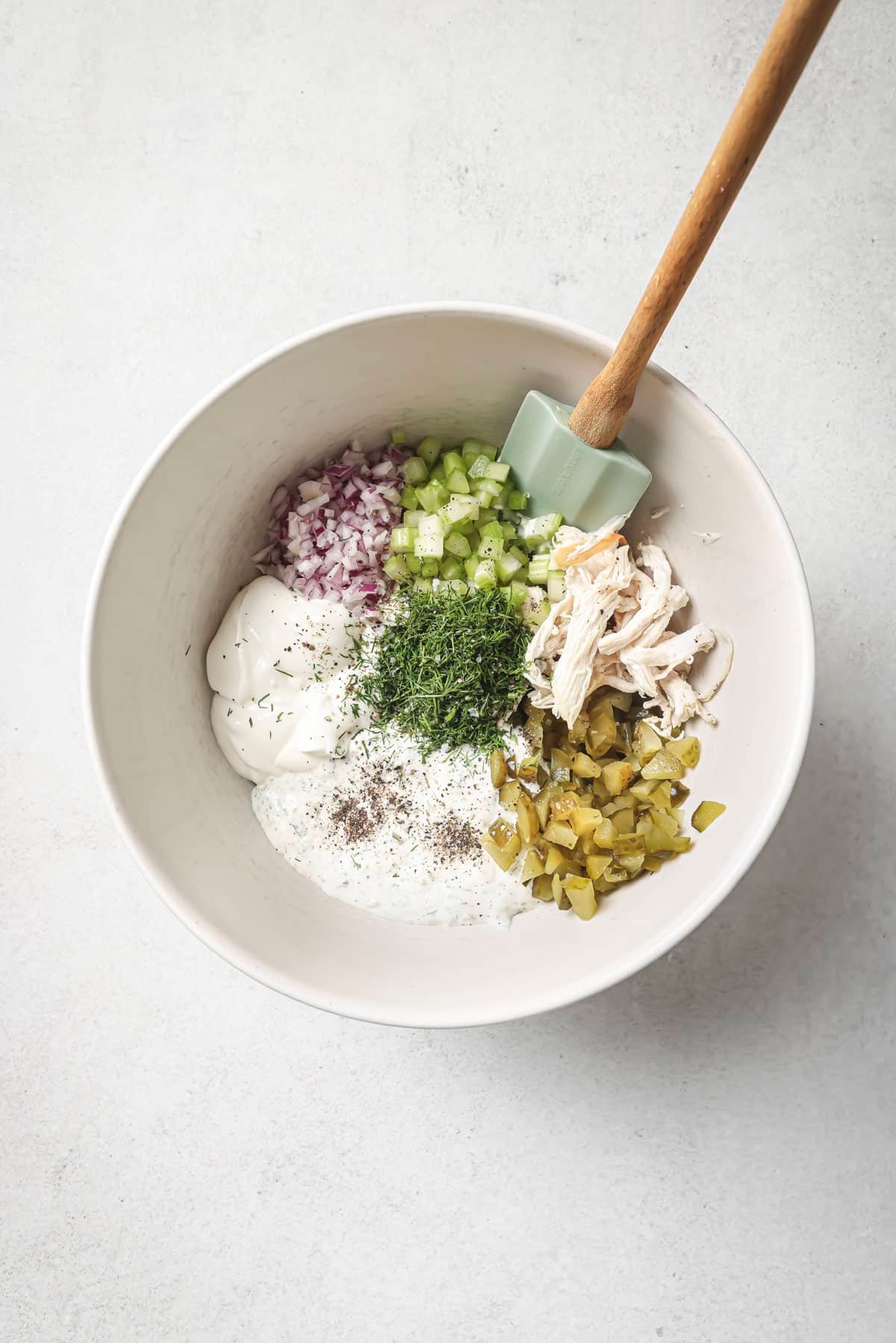 Red onion, mayo, chopped dill pickle, shredded chicken and celery in a large white bowl.