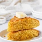 A stack of two thick slices of golden air fryer cornbread served on a white plate, topped with a pat of melting butter and honey drizzle. The texture looks moist and fluffy with crisp edges, showcasing the easy air fryer cornbread recipe.