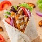 Close-up of a chicken shawarma wrap filled with air fryer chicken, fresh lettuce, tomato slices, red onion, and drizzled with creamy tahini sauce, wrapped in a soft pita and partially wrapped in parchment paper.