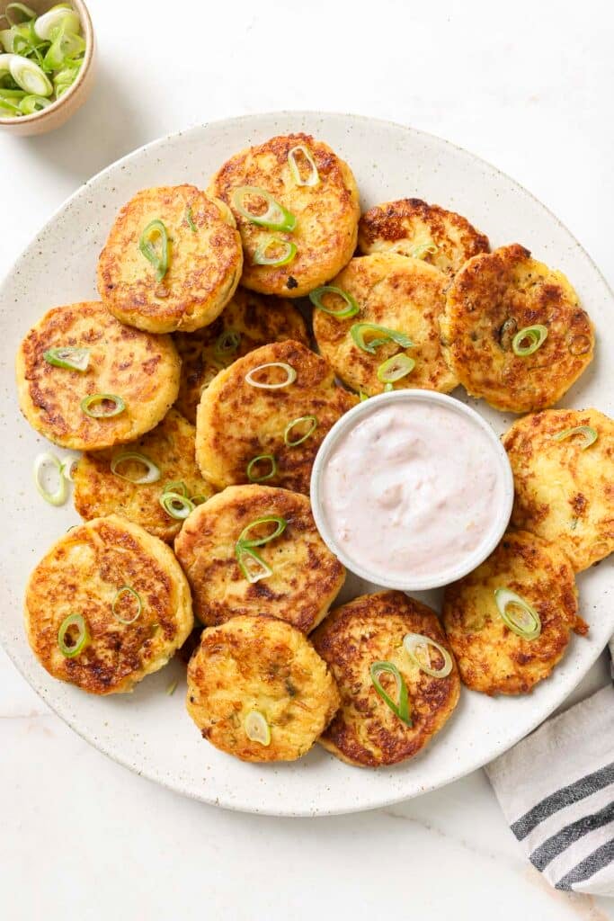 Loaded mashed potato cakes served on a large white plate with a small bowl of cranberry crème fraîche.