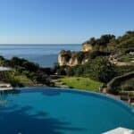Infinity pool overlooking the ocean cliffs at Vila Vita Parc Resort in the Algarve, Portugal, with white umbrellas in the foreground and clear blue sky above. Text overlay reads “the ultimate portugal itinerary – Algarve · Sintra · Lisbon.