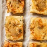 Close-up image of puff pastry apple tarts with caramelized onion, melted Gruyère cheese, and fresh thyme on parchment paper. These individual savory apple tarts are golden brown, flaky, and perfect for fall appetizers or holiday entertaining.