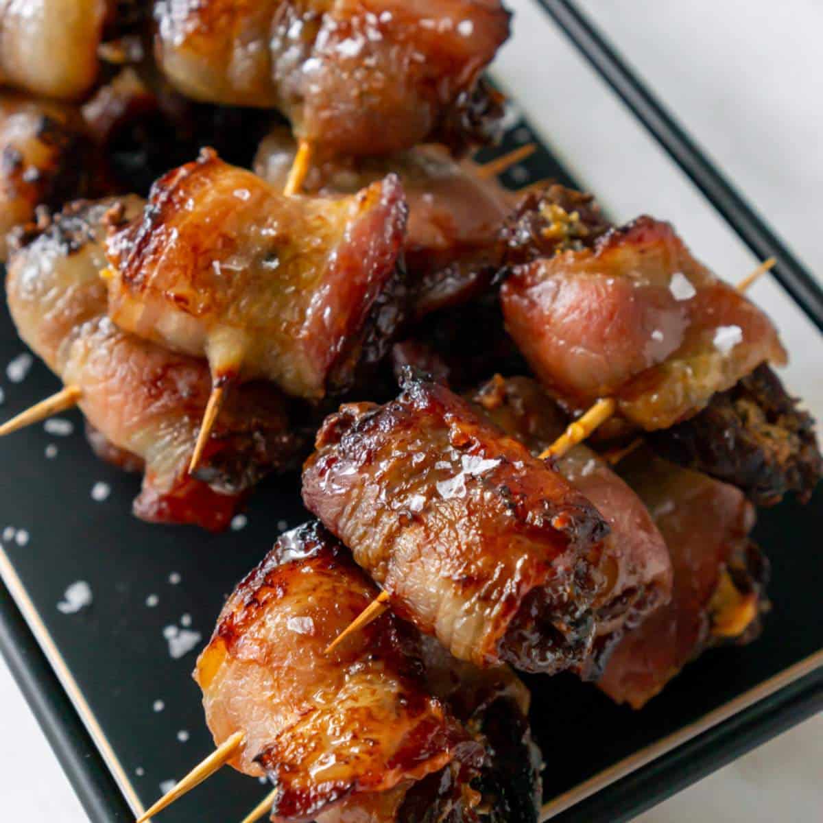 Bacon wrapped dates with toothpicks on a black plate.