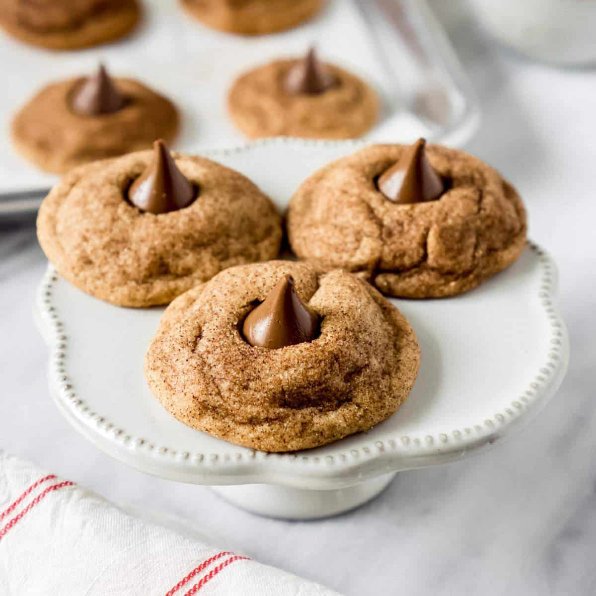 3 snickerdoodle Hershey kiss cookies on a white cake stand.
