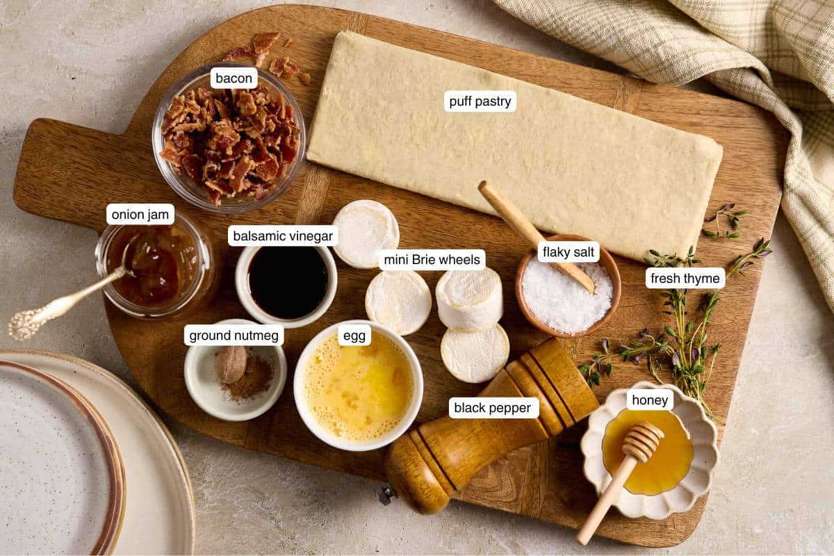 Puff pastry dough and small bowls of honey, beaten egg, flaky salt, onion jam and bacon on a wooden cutting board.