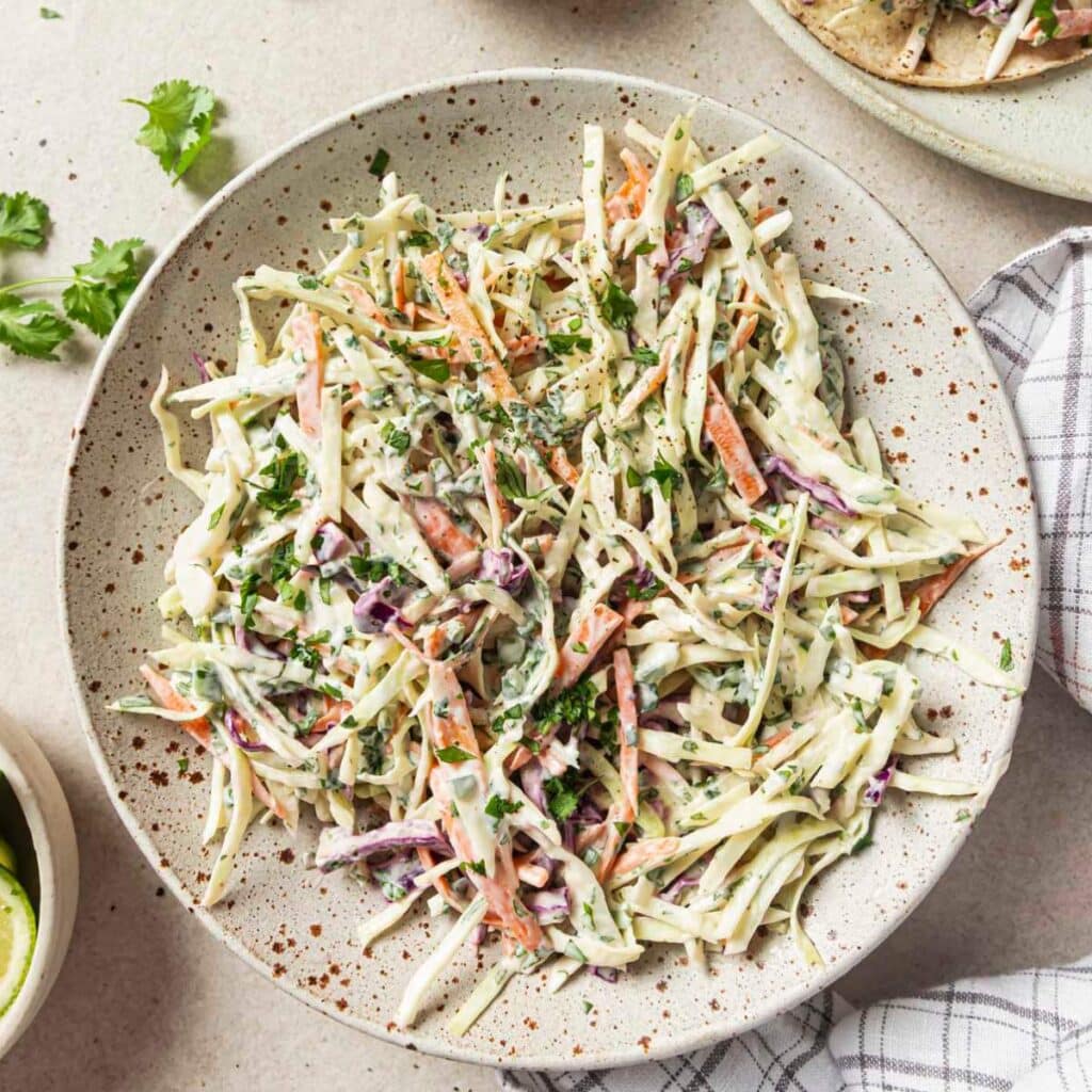 Fish taco slaw on a plate near a plate of fish tacos and freshly chopped cilantro.