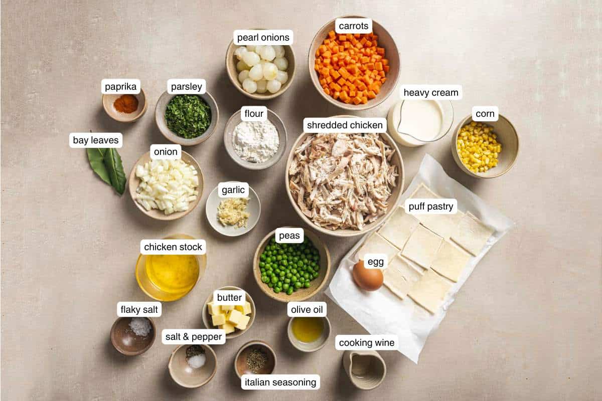 Small bowls with carrots, onions, seasonings, peas, corn, butter, flour and garlic near an egg and puff pastry dough.