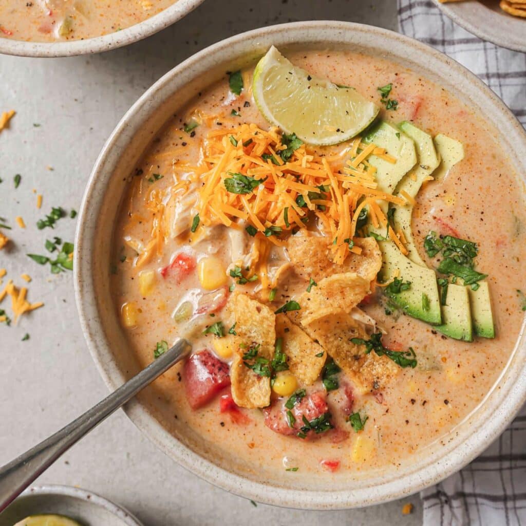 a bowl of creamy chicken fajita soup topped with tortilla chips, cilantro, and avocado in a white bowl with a spoon resting on the side