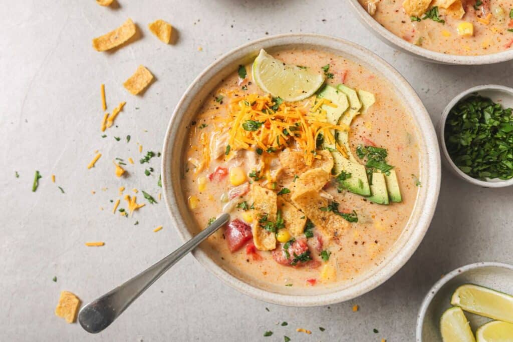 a bowl of creamy chicken fajita soup topped with tortilla chips, cilantro, and avocado in a white bowl with a spoon resting on the side