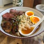 Sliced steak, jammy eggs, ramen noodles, and a spoon in a bowl.