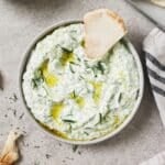 a bowl of greek tzatziki sauce topped with olive oil with a pita triangle resting inside the bowl