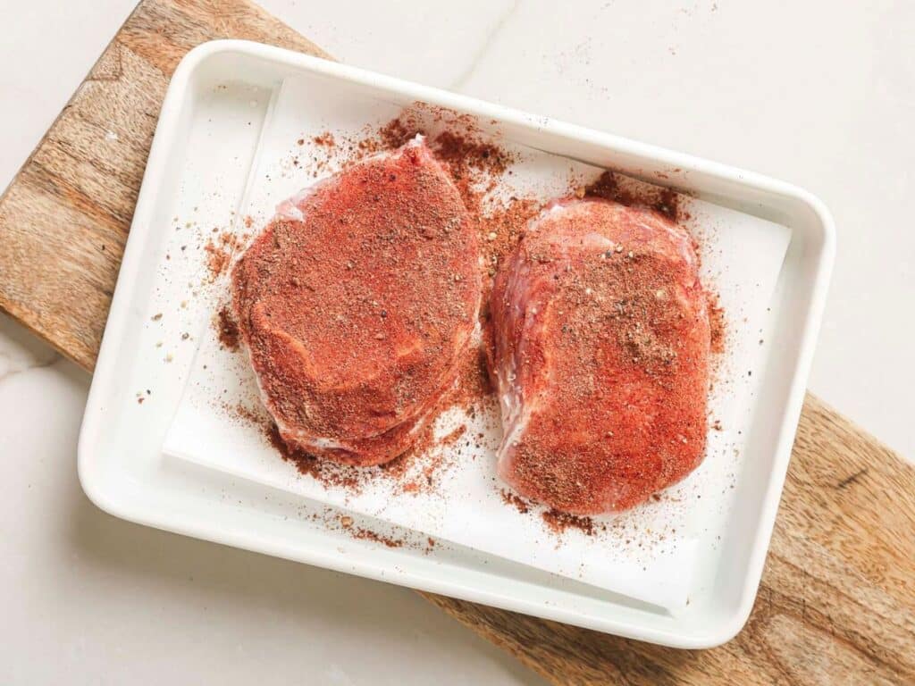 Two pork chops on a white plate covered in seasoning. 