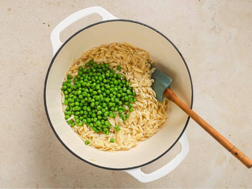 Orzo cooked in a pot, with peas resting on top and a wooden spatula on the side. 