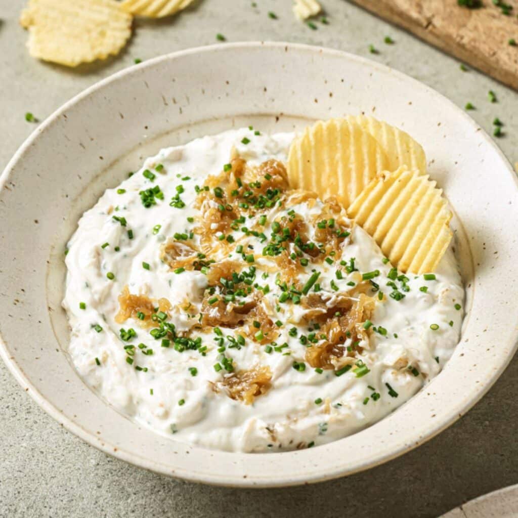 A bowl of French onion dip topped with caramelized onions and fresh chives, with two potato chips resting inside.