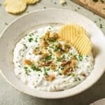 A bowl of French onion dip topped with caramelized onions and fresh chives, with two potato chips resting inside.