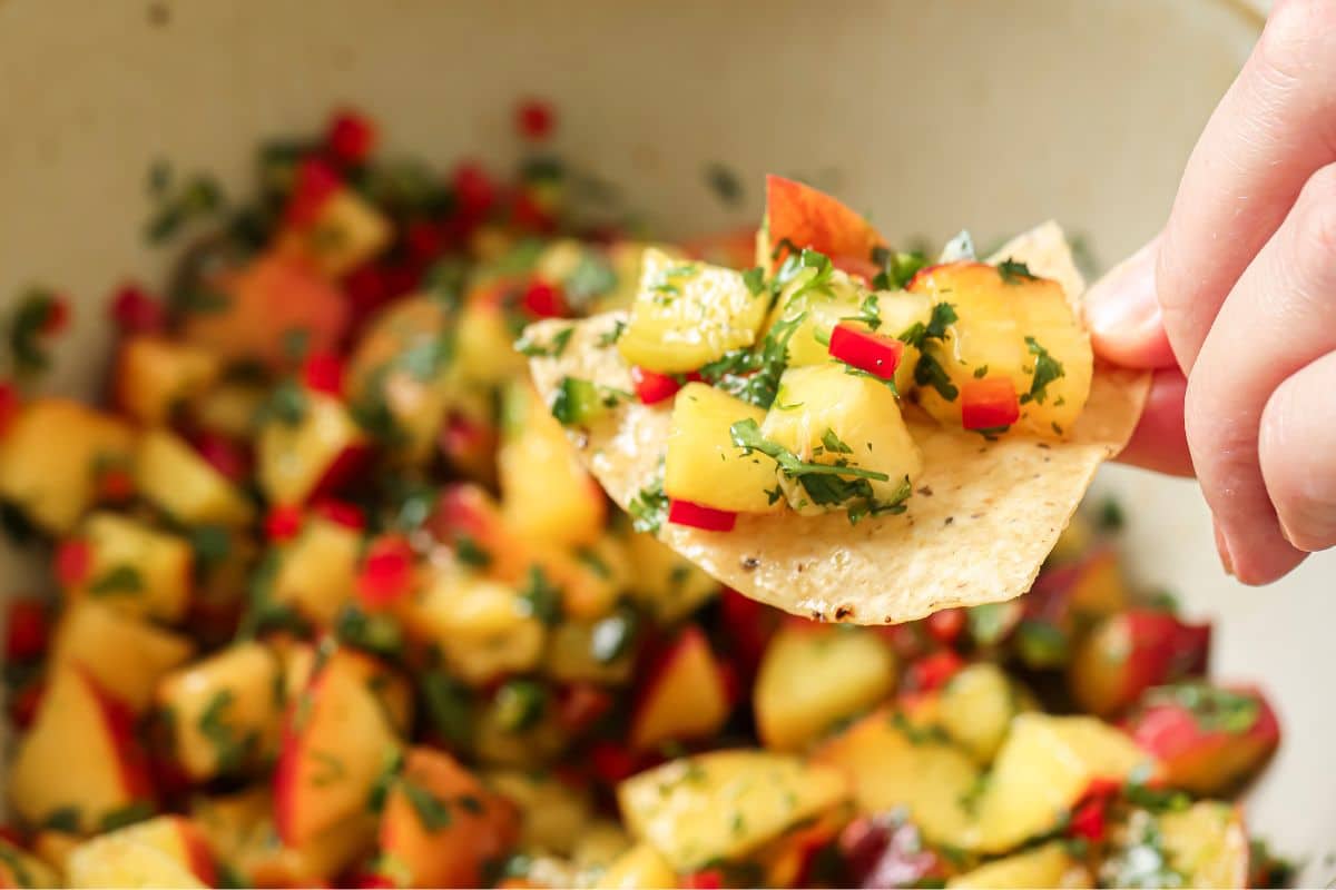A hand holding a chip with peach salsa on top of a bowl filled with peach salsa.