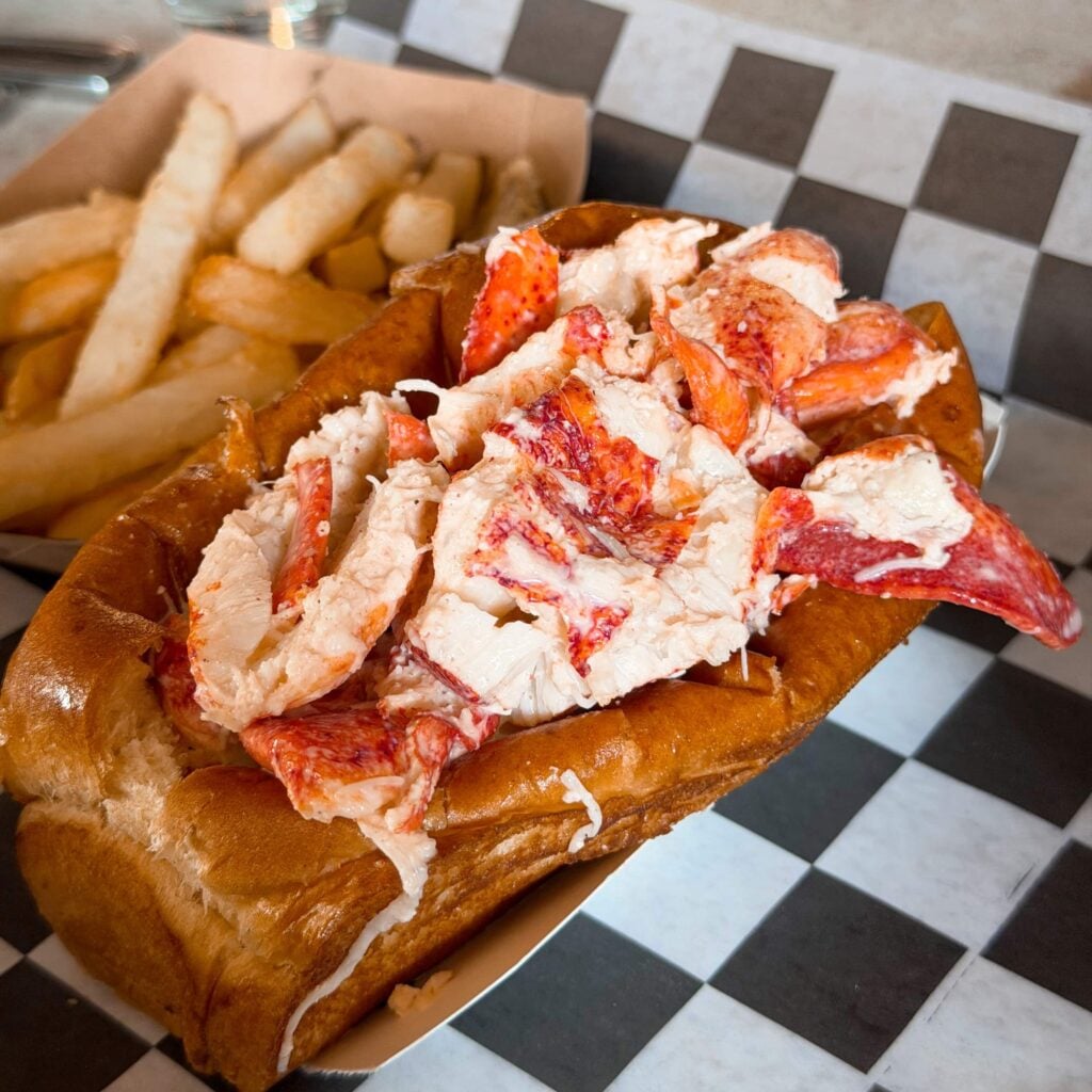 A lobster roll on a checkered piece of paper with french fries on the side.