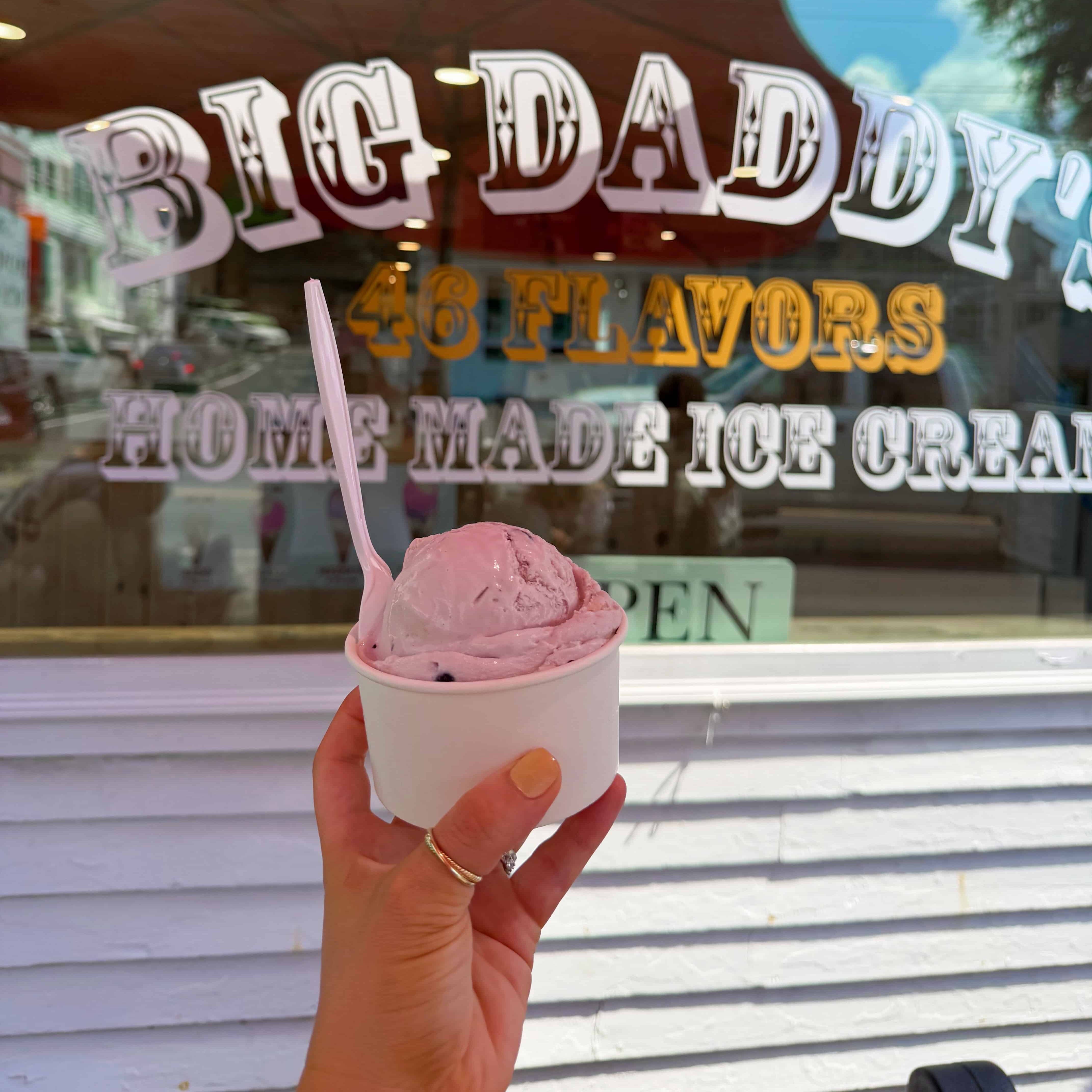 A hand holding a cup of blueberry pie ice cream in front of big daddy's ice cream shop.