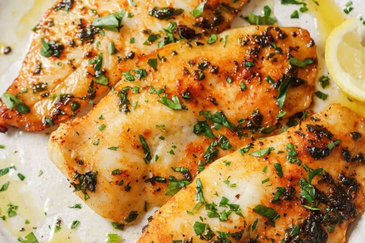 3 air fried tilapia filets topped with parsley on a white plate next to lemon wedges. 