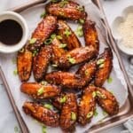 Honey garlic chicken wings on a parchment lined baking sheet topped with green onions and sesame seeds and next to a small jar of dipping sauce.