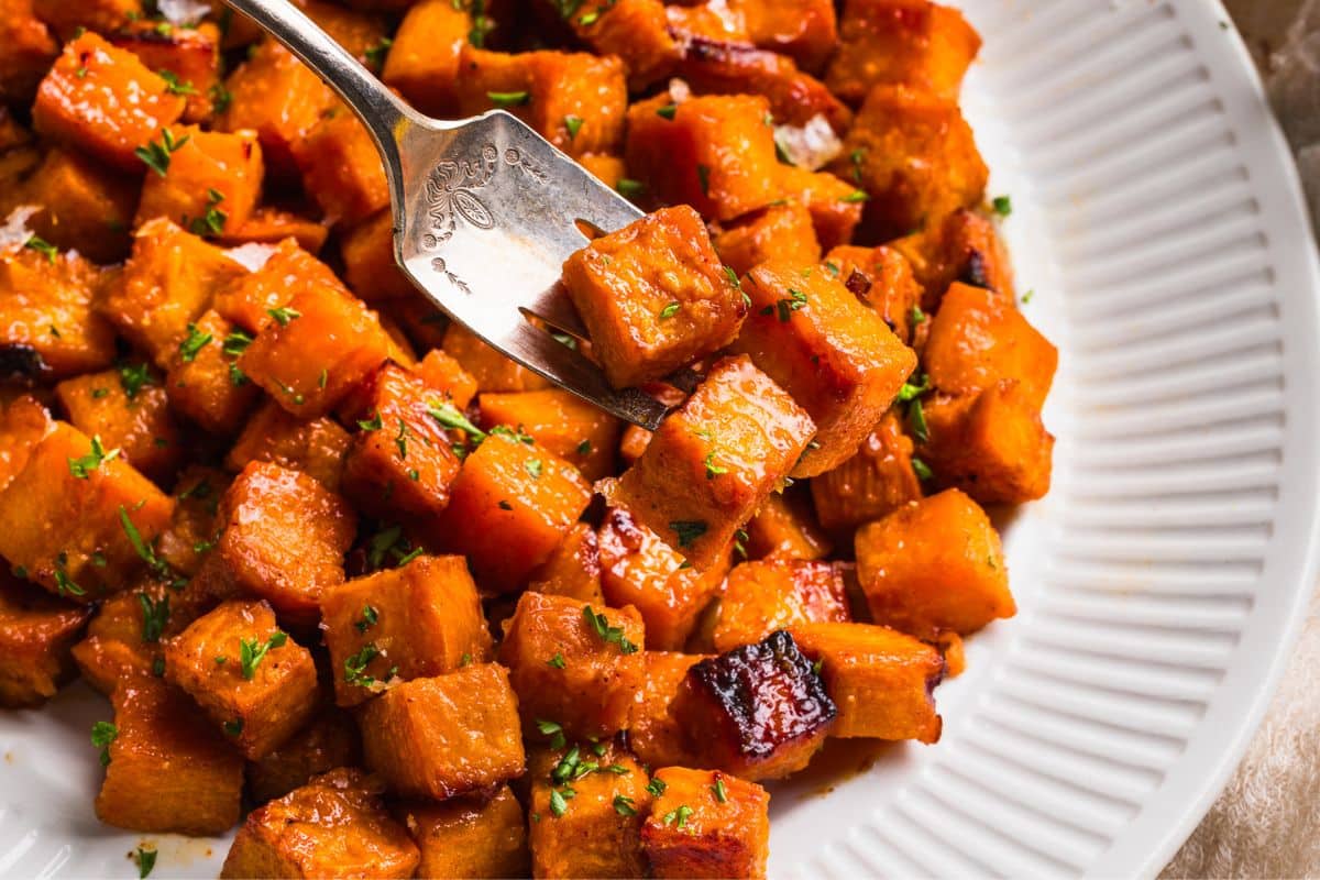 A close up shot of honey roasted sweet potatoes in a white dish with a serving fork scooping some out. 