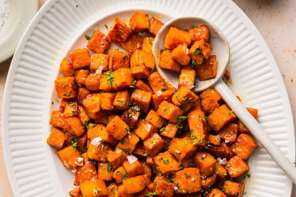 A platter filled with honey roasted sweet potatoes next to a serving spoon. 