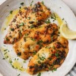 Three air fried tilapia filets topped with parsley on a white plate next to lemon wedges.