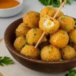 A brown bowl filled with air fryer fried goat cheese balls with one resting on top with a bite taken out to show the creamy center.