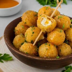 A brown bowl filled with air fryer fried goat cheese balls with one resting on top with a bite taken out to show the creamy center.