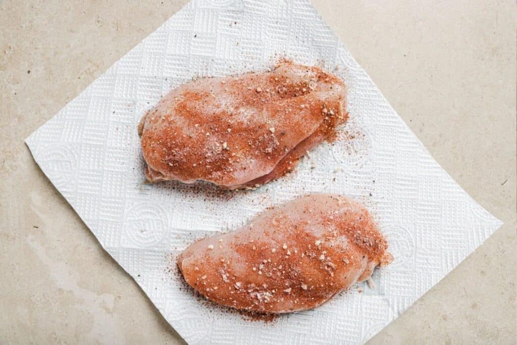 Two raw chicken breasts seasoned on top of a paper towel to absorb extra moisture. 