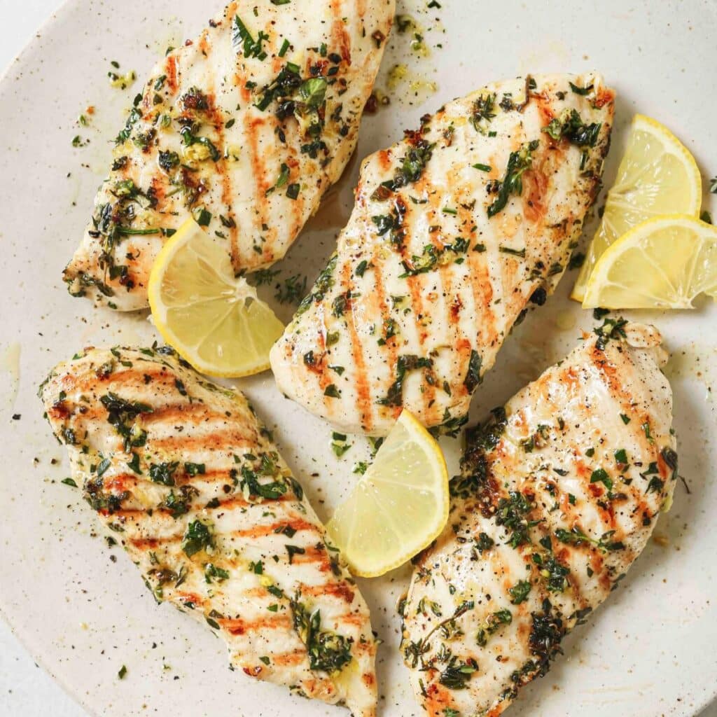 Grilled lemon herb chicken breasts on a plate with lemon wedges.